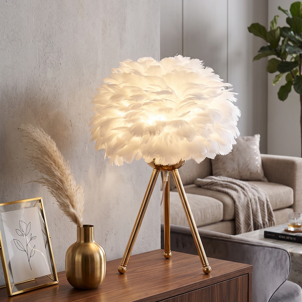 Lampe à poser avec abat-jour en plumes blanches et pied trépied en métal doré, allumée sur un meuble en bois dans un salon cosy aux tons neutres.